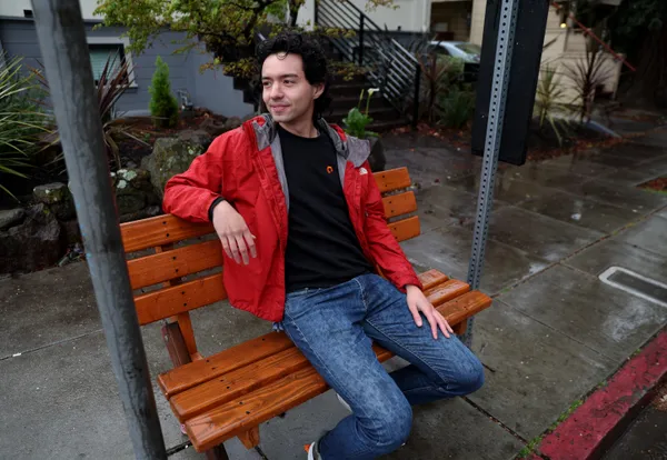 Mingwei Samuel sits on a bus stop bench that he built on the corner of Glen and Linda Avenues. He is wearing a red rain coat and jeans, and is smiling.