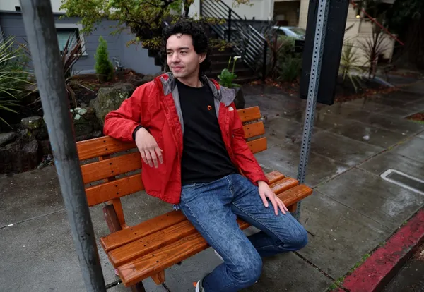 Mingwei Samuel sits on a bus stop bench that he built on the corner of Glen and Linda Avenues. He is wearing a red rain coat and jeans, and is smiling.
